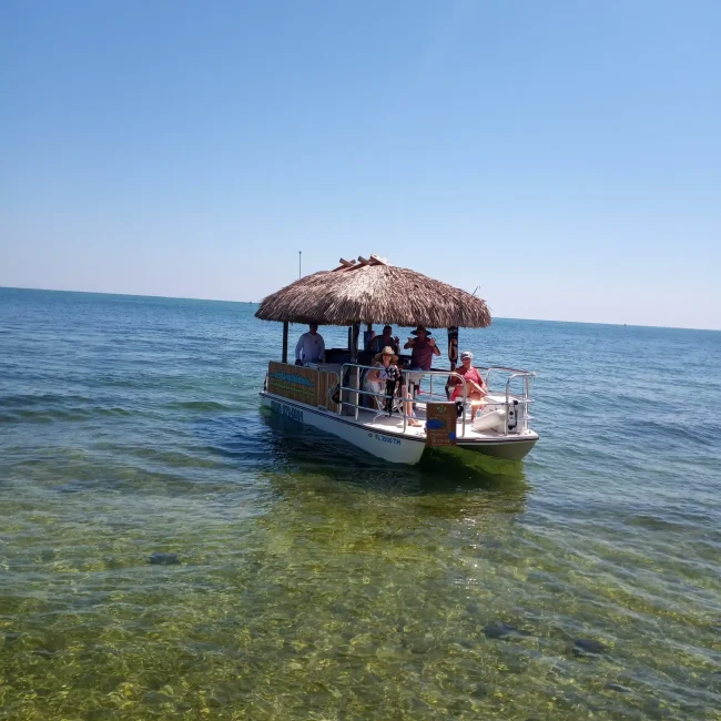 Group of people relaxing on a handcrafted tiki boats with a thatched roof, cruising on clear blue waters in the Florida Keys.