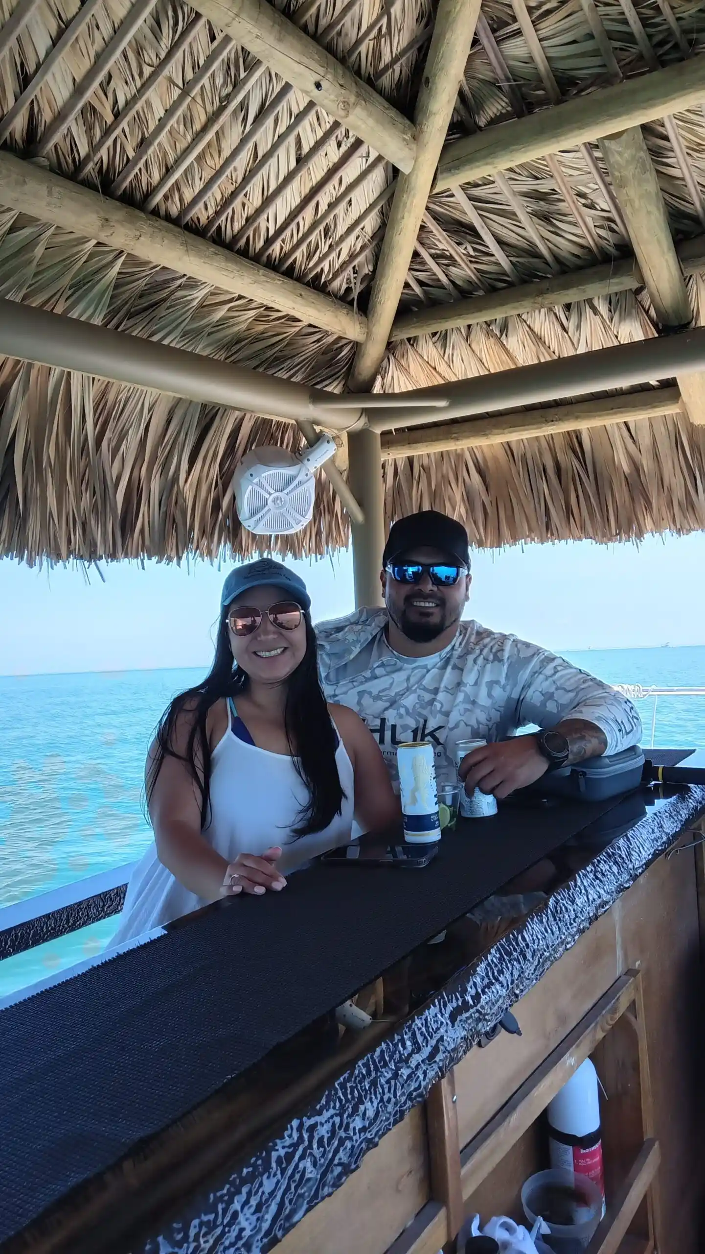 A smiling woman in a white dress and sunglasses enjoys a relaxing Key Largo tiki cruise next to a man under a thatched tiki hut, with the beautiful ocean visible.