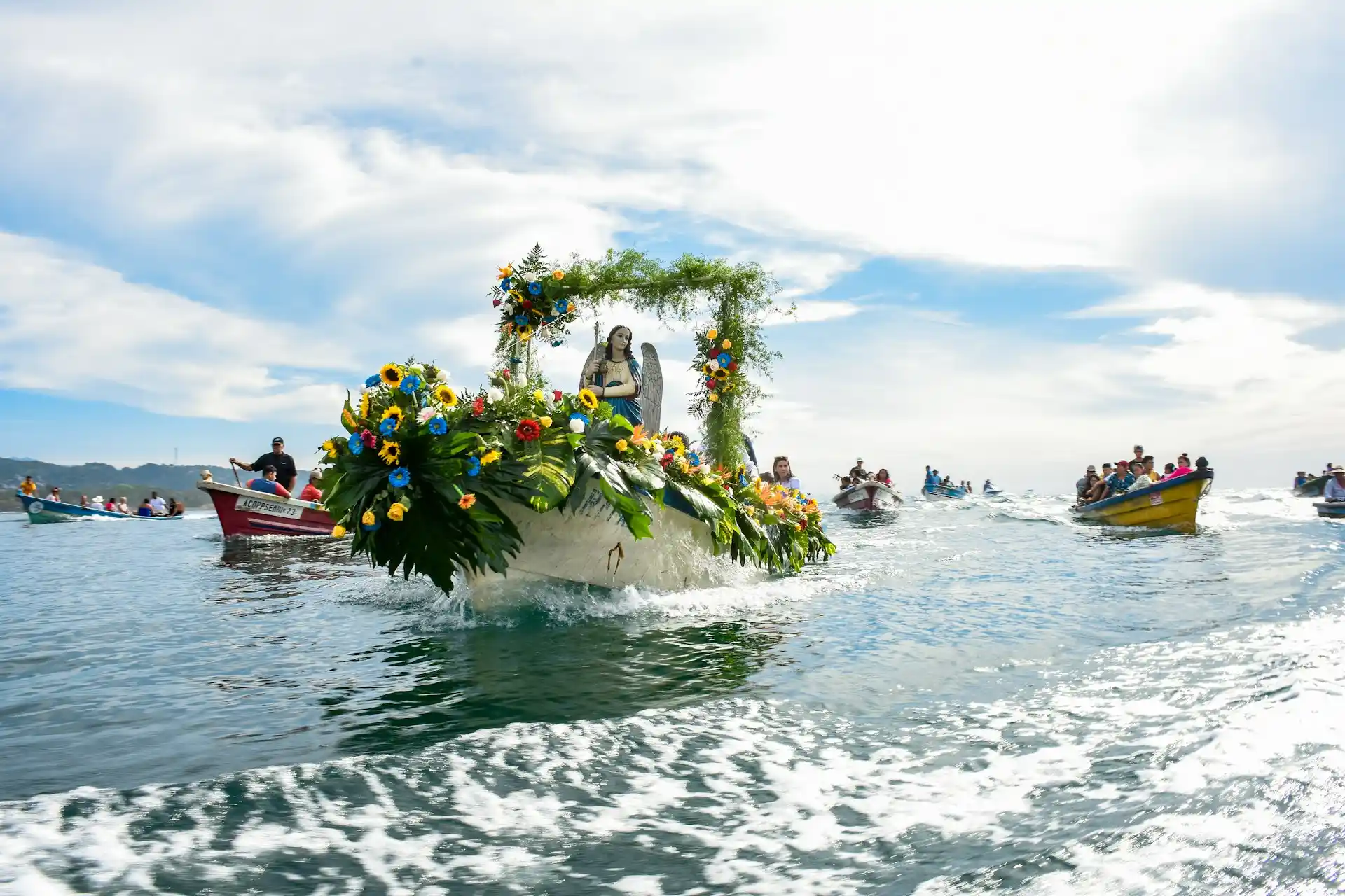 A decorated boat with a religious statue, leading a procession of smaller boats on the water under a sunny sky, indicative of unique Celebration Cruises 2025.